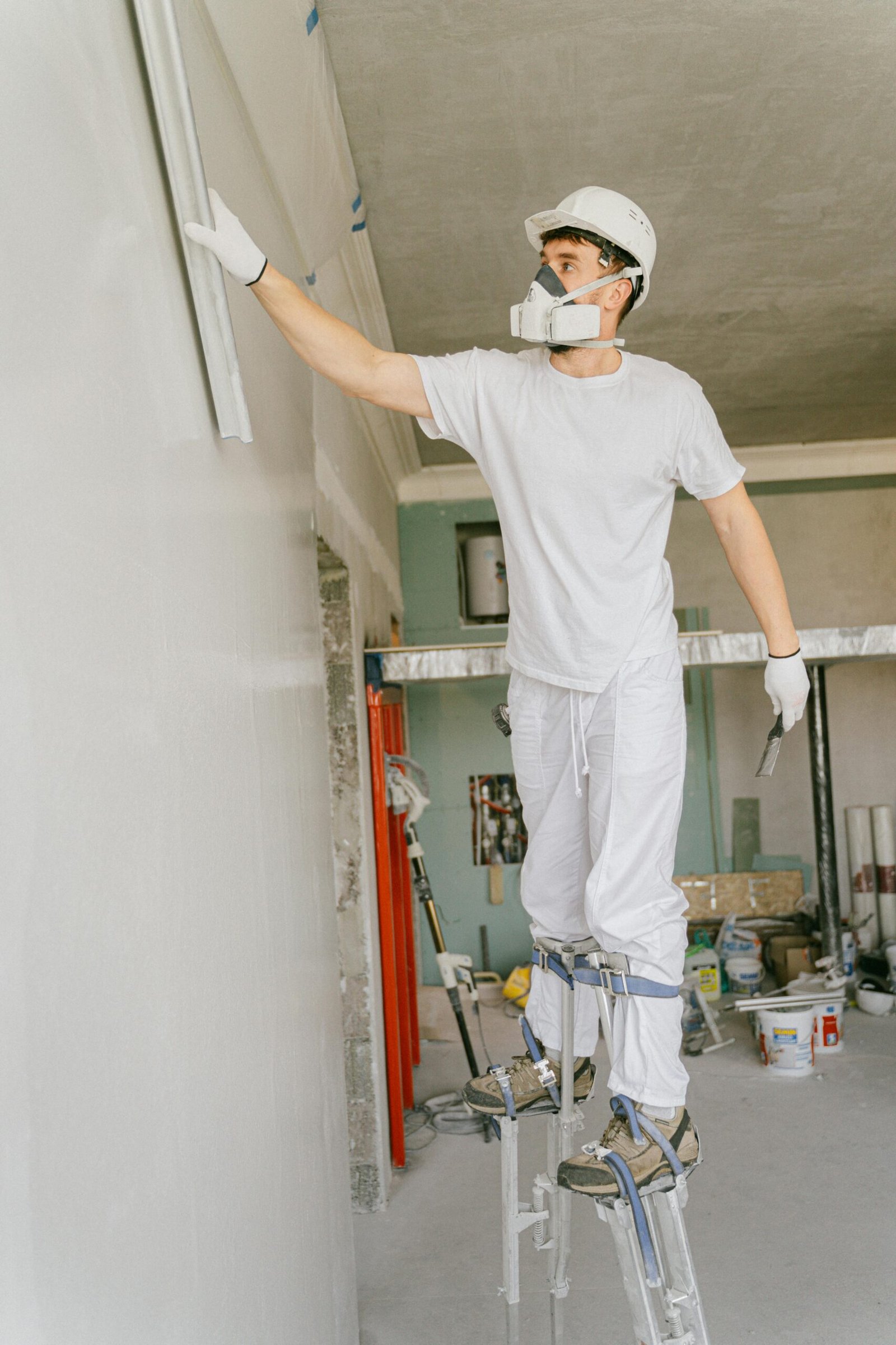 Construction worker on stilts painting a wall indoors, wearing protective gear and a hard hat.